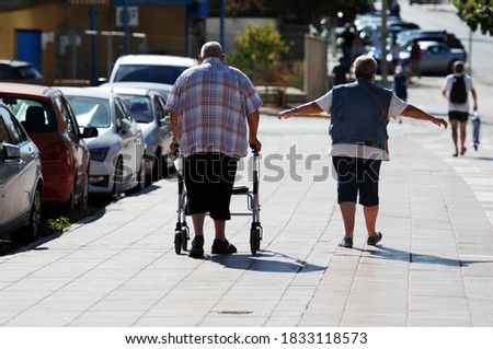 An elder couple walking on a street in Finestrat-Spain. An overweight man with a walking aid. An older woman spreads his arms.