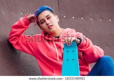 Stylish young woman with short colored hair sitting with her blue plastic skateboard in skatepark. Youth concept.