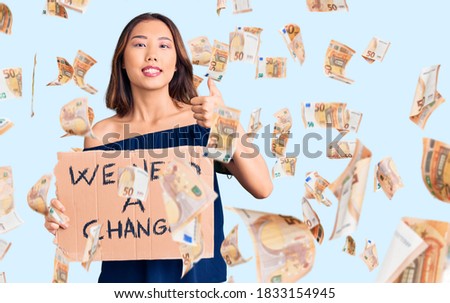 Young beautiful chinese girl holding we need a change banner smiling happy and positive, thumb up doing excellent and approval sign