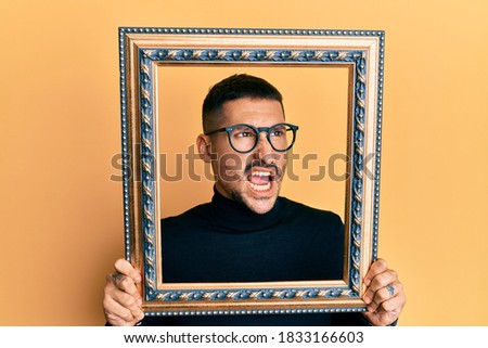 Handsome man with tattoos holding empty frame angry and mad screaming frustrated and furious, shouting with anger. rage and aggressive concept. 