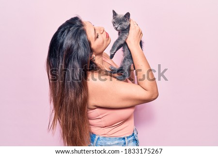 Young beautiful latin woman smilling happy. Standing with smile on face holding adorable cat over isolated pink background