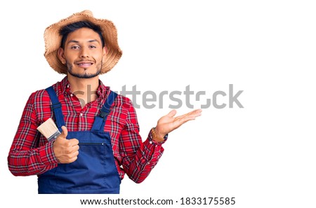 Handsome latin american young man weaing handyman uniform showing palm hand and doing ok gesture with thumbs up, smiling happy and cheerful 