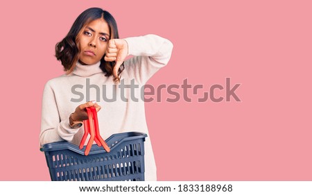 Young beautiful mixed race woman holding supermarket shopping basket with angry face, negative sign showing dislike with thumbs down, rejection concept 