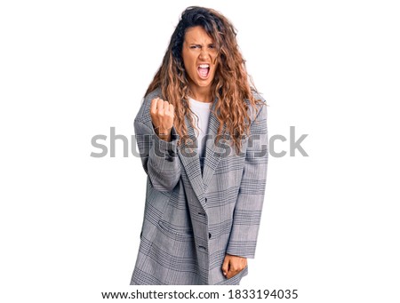 Young hispanic woman with tattoo wearing business oversize jacket angry and mad raising fist frustrated and furious while shouting with anger. rage and aggressive concept. 