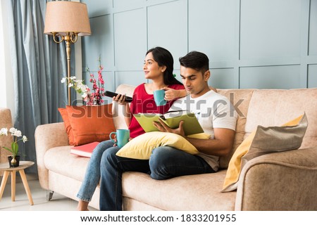Indian man reading book and wife watching tv while sitting on sofa or couch