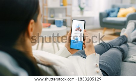 Over the Shoulder Shot of a Hungry Young Woman Sitting on a Couch at Home and Using Smartphone for Ordering a Food via Advanced Modern Food Delivery App. She is Waiting for Delivery. Royalty-Free Stock Photo #1833205129
