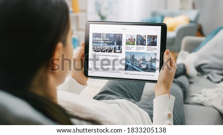 Over the Shoulder Shot of a Young Woman at Home Lying on a Couch and Using Tablet Computer in Horizontal Landscape Mode for Reading News about Technology. Girl Using Touchscreen Device. Royalty-Free Stock Photo #1833205138