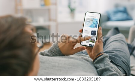 Young Man at Home is Lying on a Coach and Using Smartphone for Scrolling and Reading News about Technological Breakthroughs. He's Sitting On a Couch in His Cozy Living Room. Over the Shoulder Shot Royalty-Free Stock Photo #1833205189