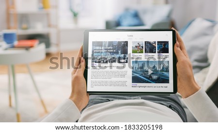 Young Woman at Home Lying on a Couch Using Tablet Computer in Horizontal Landscape Mode for Reading Technology News. Girl Using Touchscreen Device, Browsing Internet, Blogs. POV Shot. Royalty-Free Stock Photo #1833205198