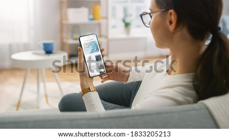 Young Woman at Home Uses Smartphone for Scrolling and Reading News about Technological Breakthroughs. She's Sitting On a Couch in Her Cozy Living Room. Over the Shoulder Shot Royalty-Free Stock Photo #1833205213