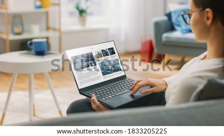 Young Woman at Home Is Using Laptop Computer for Scrolling and Reading News about Technological Breakthroughs. She's Sitting On a Couch in His Cozy Living Room. Over the Shoulder Shot Royalty-Free Stock Photo #1833205225