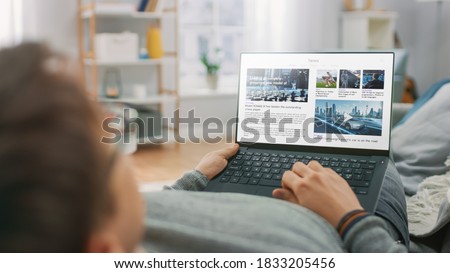 Young Man at Home Laying on a Couch and Using Laptop Computer for Reading News about Technological Breakthroughs. Guy is Using Laptop Device, Browsing Internet, Watching Content, Videos. Royalty-Free Stock Photo #1833205456