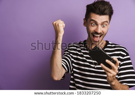 Photo of excited man making winner gesture while playing game on cellphone isolated over purple background