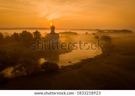 Aerial shot of a Russian Orthodox church above a misty field lit by the rising sun. A pond near the church, a river in the background. No people.