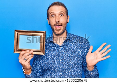 Young handsome man holding empty frame celebrating achievement with happy smile and winner expression with raised hand 