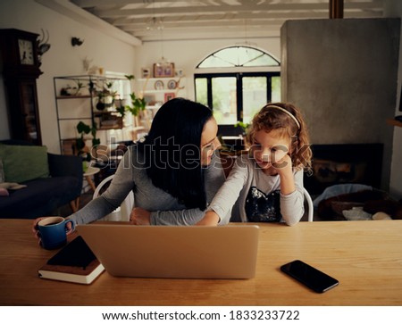 Cheerful woman looking at preschool daughter pointing at laptop screen at home