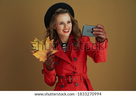 Hello november. smiling 40 years old woman in red coat and black beret with leather gloves and yellow autumn maple leaves taking selfie with mobile phone on brown background.