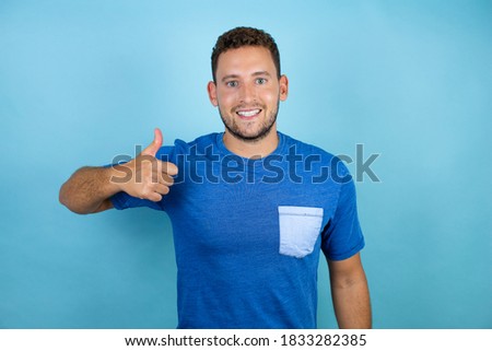 Young handsome man wearing blue casual t-shirt over isolated blue background doing happy thumbs up gesture with hand. Approving expression looking at the camera with showing success
