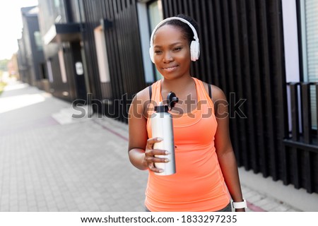 fitness, sport and healthy lifestyle concept - young african american woman in headphones drinking water from bottle outdoors