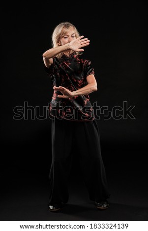 Mature blonde woman practicing Tai Chin Chen style in a black background wearing a traticional chinese black jacket with red decoration, black trousers and traditional shoes with ying yang symbol