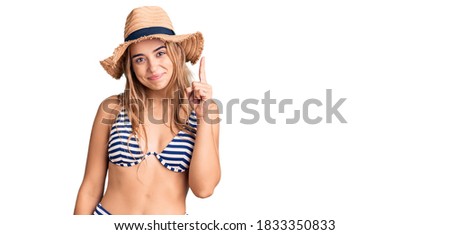 Young beautiful blonde woman wearing bikini and hat showing and pointing up with finger number one while smiling confident and happy. 