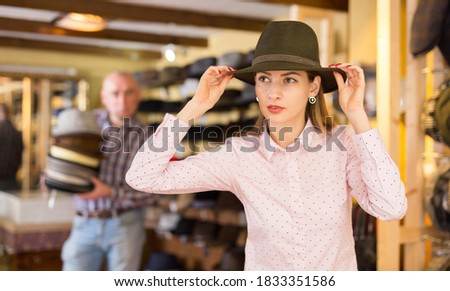 Young stylish woman fitting on classic hat in headwear shop, male seller holding stacked hats