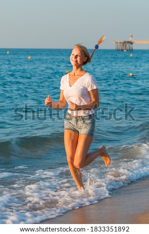 Smiling adult woman in white T-shirt is jogging on the beach near the ocean.