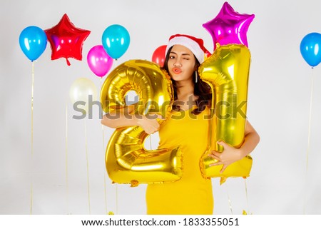 Happy, a charming, long curly-haired Asian woman holding two golden balloons, making twenty-one numbers, delighted with her age that matches her birthdays and Christmas.