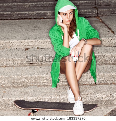 beautiful young girl in green hoodie talks on cellphone sitting on stone stairs with her legs on skateboard