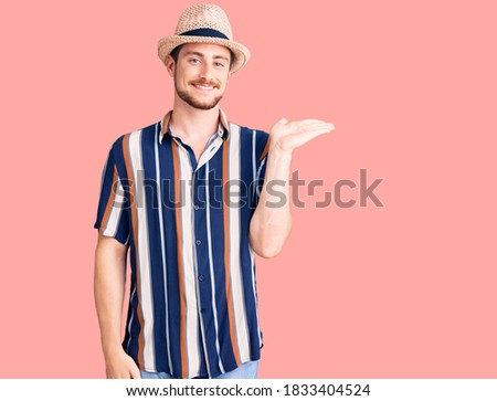 Young handsome caucasian man wearing summer hat smiling cheerful presenting and pointing with palm of hand looking at the camera. 