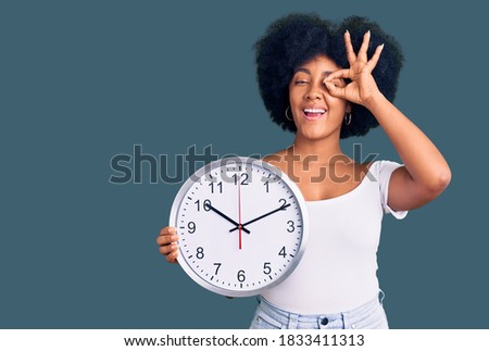 Young african american girl holding big clock smiling happy doing ok sign with hand on eye looking through fingers 