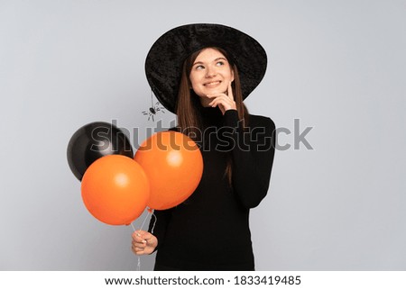 Young witch holding black and orange air balloons thinking an idea while looking up