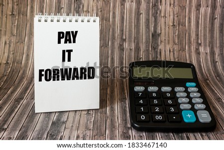 The phrase Pay It Forward with text on a notepad, placed on a wooden board, next to a calculator.