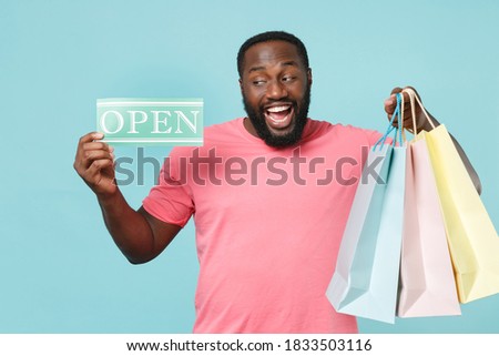 Excited young african american man in casual pink t-shirt isolated on blue background studio portrait People lifestyle concept. Hold package bag with purchases after shopping sign card with OPEN title