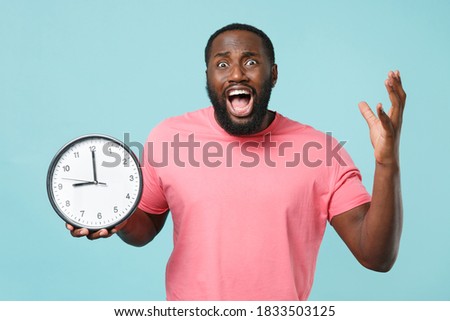 Worried young african american man 20s in casual pink t-shirt isolated on blue background studio portrait. People emotions lifestyle concept. Mock up copy space. Hold clock, screaming spreading hands