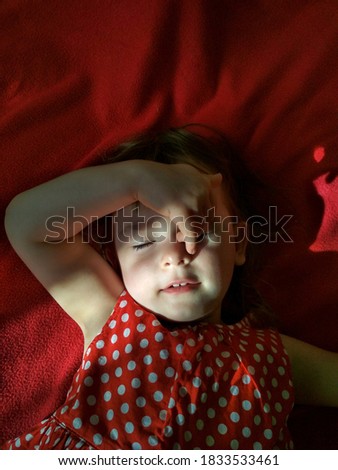 Portrait of a girl with closed eyes, who lies on a red blanket. The girl is wearing a red polka-dot dress. Top view.