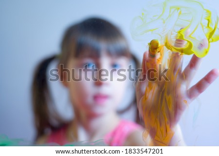 Beautiful young girl painting painting artwork on glass with colorful hands and finger. Happy childhood, art, painting lessons concept. Selective focus on finger. Horizontal image.
