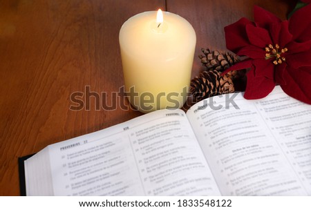 A bible on the table in the light of a candle with pine cones and Christmas poinsettia flowers.
