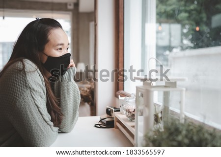 Asian woman wearing fabric handmade black cotton protective mask sitting and looking through the window with negative space. Coronavirus mask wearing trends.