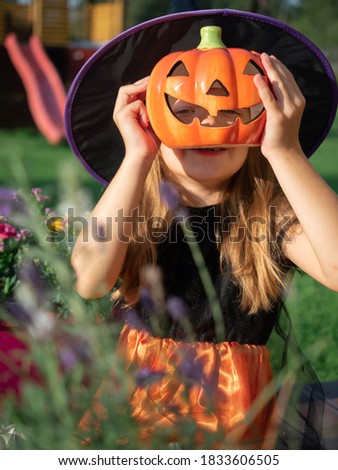 6-7 years old adorable girl in witch costume hiding behind artificial pumpkin for home decor 
