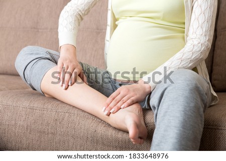 Leg cramps during pregnancy. Closeup of hands massaging swollen foot while sitting on sofa. Royalty-Free Stock Photo #1833648976