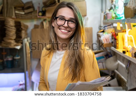 Successful small business owner in casual wearing yellow cardigan, standing in her store, looking at camera. Woman in spectacles smiling, holding stack of paper documents, store on background. 