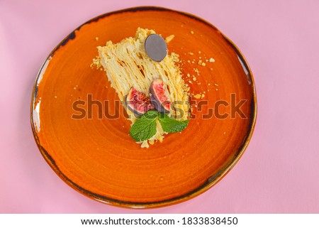 One piece of Napoleon cake with fresh fig fruit and a piece of chocolate served on a bright orange plate over pastel pink background. Delicious dessert for tea or coffee time. Close up.