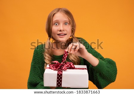 Image of charming blonde girl 12-14 years old in warm green sweater holding present box with red bow. Studio shot, yellow background, isolated. New Year Women's Day birthday holiday concept