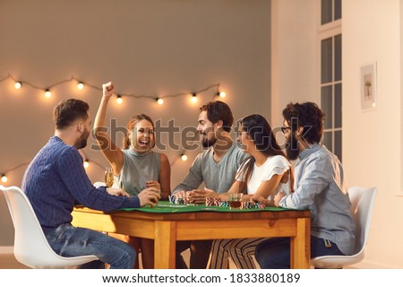 Overjoyed, happy young woman laughing and raising arm, celebrating her victory in game of poker, sitting at table with friends at casino themed party at home or club
