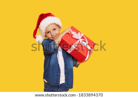 Portrait of a satisfied little child boy in christmas Santa hat. laughing isolated over yellow background. Holds a gift box. Preparing for the New Year holidays Royalty-Free Stock Photo #1833894370