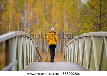 Young woman in yellow jacket slowly walking on bridge at natural park in colorful autumn day. Spending time alone in nature. Peaceful atmosphere. Back view. 