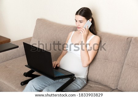 Pretty pregnant woman talking on cellphone and using laptop while sitting on sofa at home.