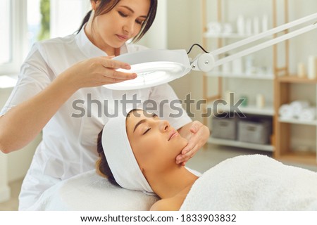 Woman cosmetologist looking at serene young womans face anc checking skin condition during skin examination under lamp in beauty salon. Professional skin checking concept Royalty-Free Stock Photo #1833903832
