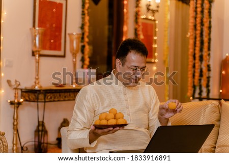 Young Man in tradional wear at home with laptop and ladoo plate in hand Royalty-Free Stock Photo #1833916891